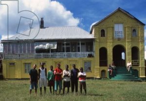 A picture of the ramble house painted in yellow with green steps and people standing in front and sitting on the steps.